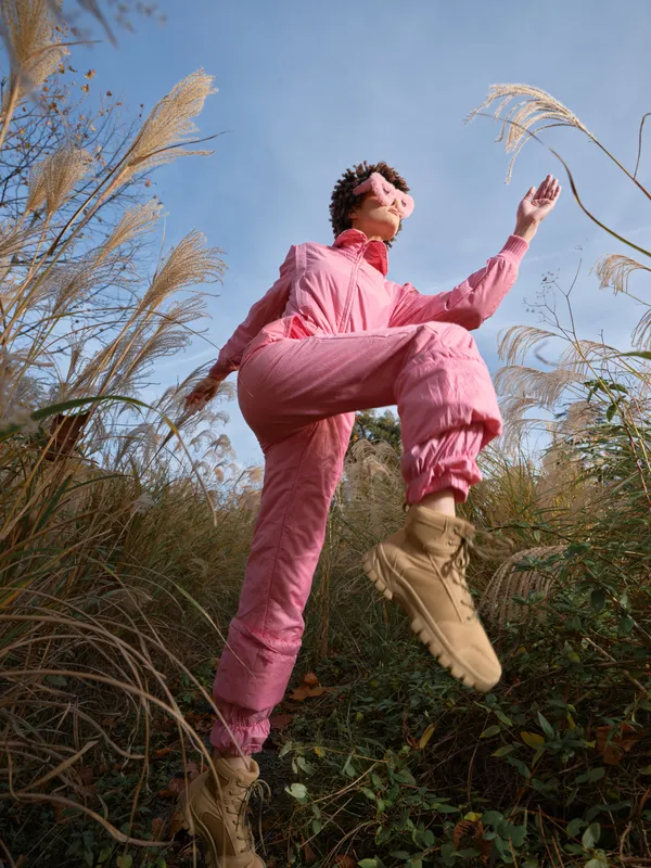 Outdoor scene with a woman leaping in a pink outfit among tall grasses, blue sky above, capturing energy, movement and carefree joy in nature, a vibrant moment of bold leap and freedom