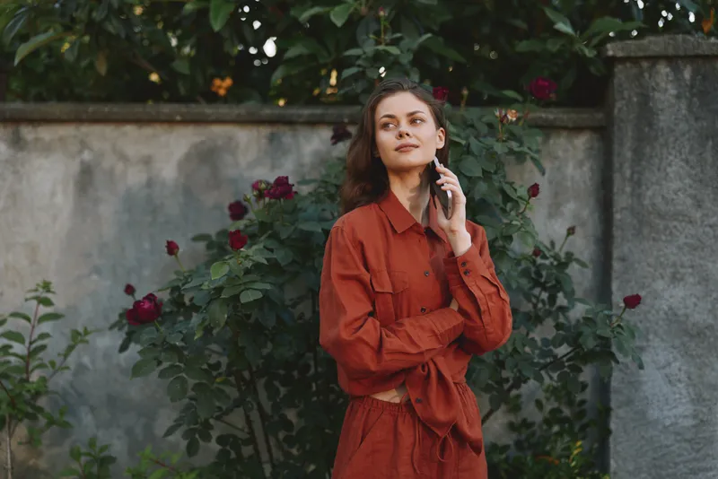 Smiling woman in a rust red dress poses outdoors among blooming roses, applying makeup with a soft background. Concept of beauty, wellness, and self care in a warm natural setting