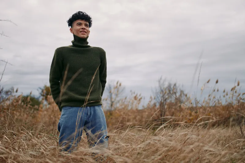 Young man stands in tall field grass wearing a turtleneck sweater, captured in a candid, authentic moment that emphasizes genuine presence, natural posture, and an honest, grounded emotional tone.