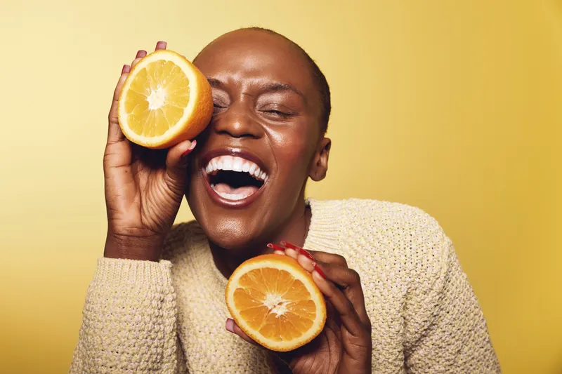 Happy African woman with dark skin holds two orange halves near face laughing joyfully on yellow background. Concept of health, vitamin, fresh fruit, and natural beauty with vibrant mood.