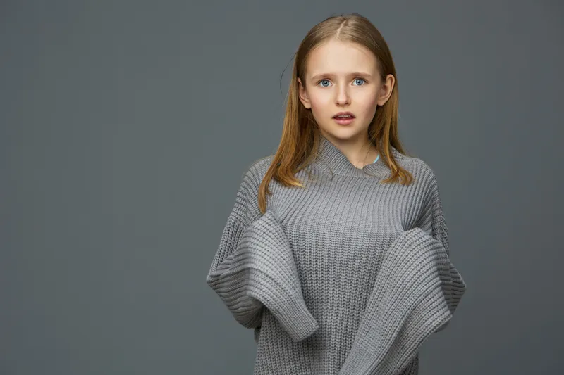 Young girl in a cozy gray sweater, expressing surprise against a neutral gray background, perfect for winter lifestyle concepts and emotional portraits