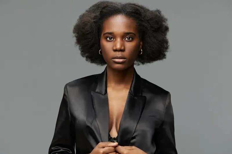 Confident African woman in a sleek black blazer, showcasing natural beauty and strength against a neutral gray background