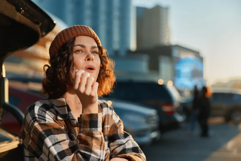 Woman portrait urban candid lifestyle scene at golden hour, beanie and plaid jacket, thoughtful expression by parked cars, authenticity, mindful living and emotional storytelling with golden hour glow