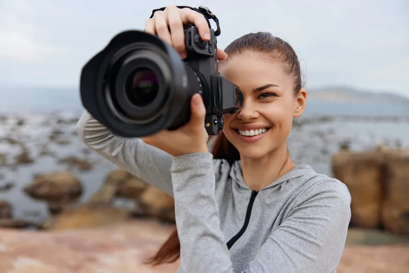Smiling young woman with a camera in casual sportswear captures nature on a rocky beach, showcasing photography passion and joy in a serene outdoor setting