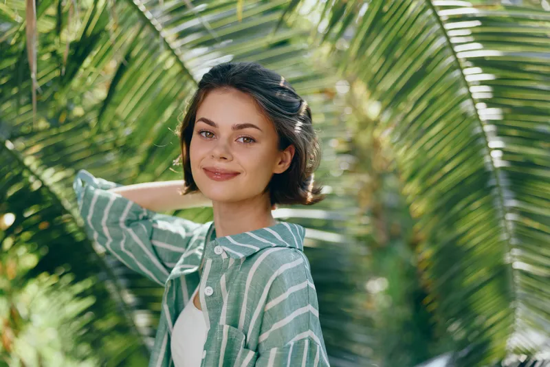 A cheerful woman stands outdoors among lush palm leaves, wearing a striped shirt and a light smile, exuding relaxed confidence in a sunny tropical setting.