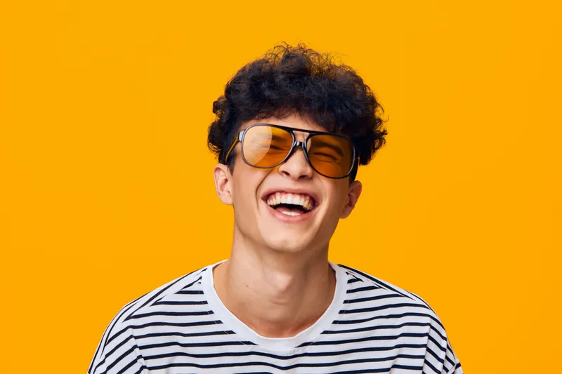 young man laughing joyfully wearing orange sunglasses and striped t-shirt on vibrant yellow background, expressing happiness and carefree attitude