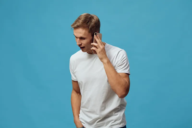 Man talking on mobile phone wearing casual white t-shirt against blue background. Studio portrait of a young adult male, expressing communication and technology, isolated on plain vibrant backdrop, li