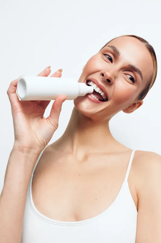 Young woman with clean skin using white cosmetic bottle on face, smiling, skincare routine, beauty concept, bright background, natural makeup, casual outfit, fresh look