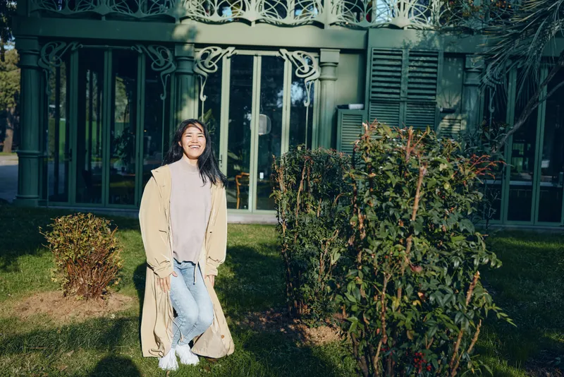 Woman in a long beige coat stands in a garden setting, casual denim and white sneakers, sunny afternoon, greenery and fence backdrop, relaxed lifestyle photo