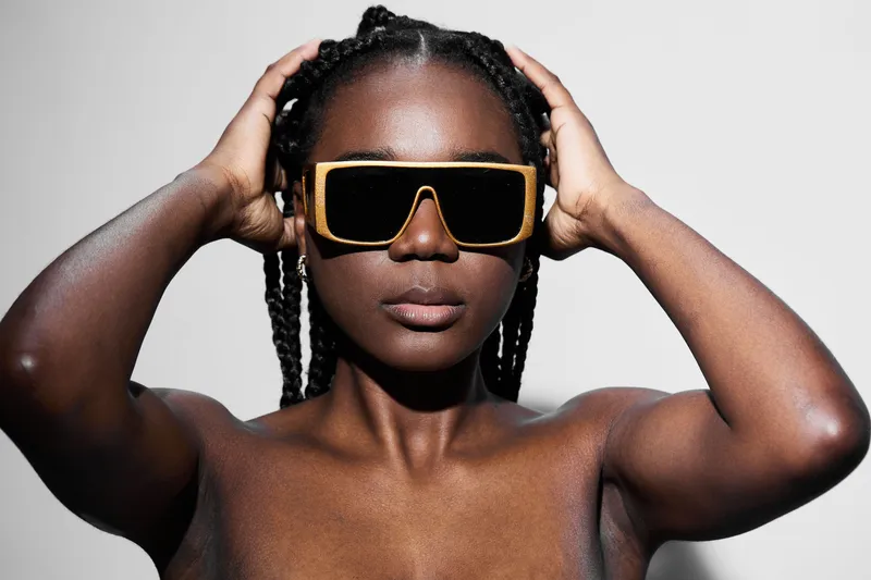 Confident Black model with braided hair wearing oversized sunglasses, showcasing a bold and modern style against a minimalist background