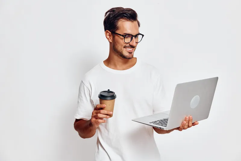 Smiling young man wearing glasses holds a laptop while enjoying coffee, looking at the screen with a cheerful expression against a solid white background.
