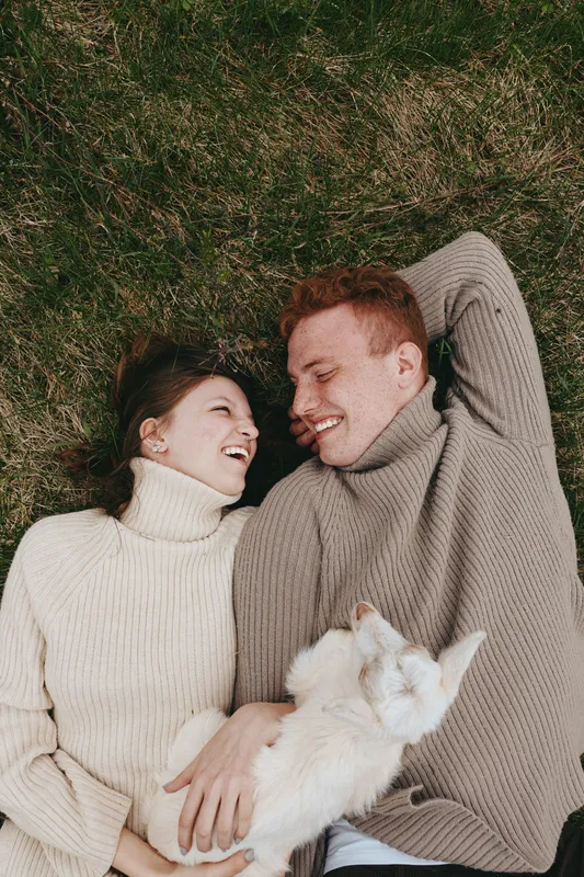 A couple lies on grass in cozy knitwear, smiling as a goat kid cuddles between them, capturing a warm lifestyle moment of affection outdoors bonding and gentle countryside play