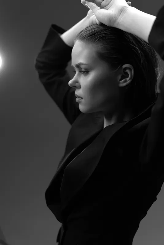 Black and white portrait of a serious woman in a dark jacket with hands raised, showing strong emotion and elegant style.