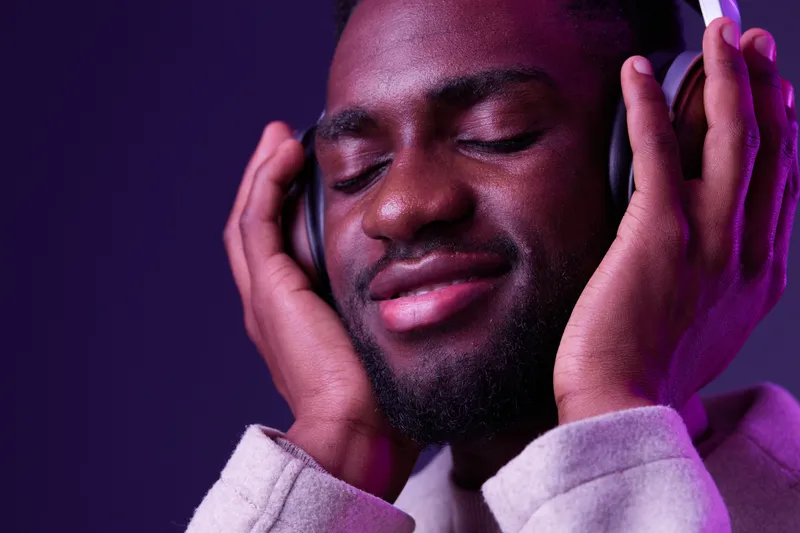 Young Black man enjoying music with headphones, eyes closed, calm expression, purple background, contemporary style, perfect for ads about relaxation and mindfulness