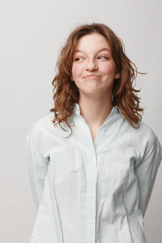 Young woman with curly hair wearing white shirt looks sideways with playful smile. Studio portrait on light background. Concept of casual style, youth, and positive emotions.