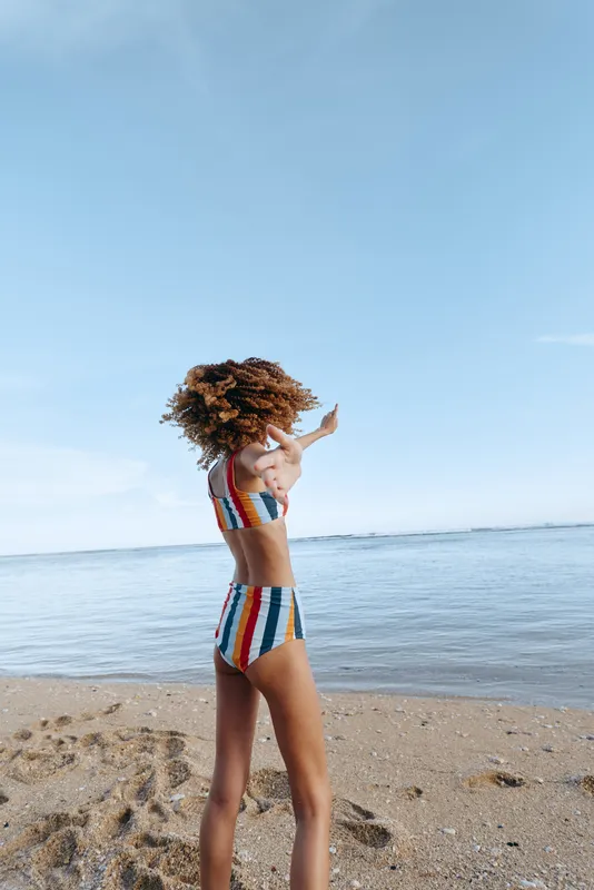 Beach child girl on sandy shore, holding curly hair, wearing striped swimsuit, ocean horizon and clear sky behind, summer sunlight, calm vacation mood, standing outdoors, bright colors