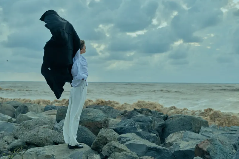 Cloaked figure on a rocky shoreline faces the sea as wind sweeps a flowing garment, creating a dramatic silhouette under cloudy skies, evoking mystery and resilience