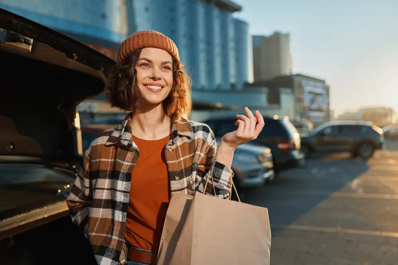 Woman shopping with bag, smiling in urban parking lot at golden hour glow, candid lifestyle portrait showing authenticity, mindful living and emotional storytelling in city scene