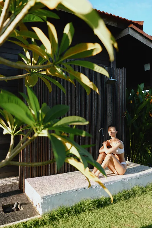 Lifestyle scene of a woman outdoor relaxation by a wooden wall, sitting on a low ledge, wearing casual clothing, serene expression, tropical garden surroundings under bright sun.