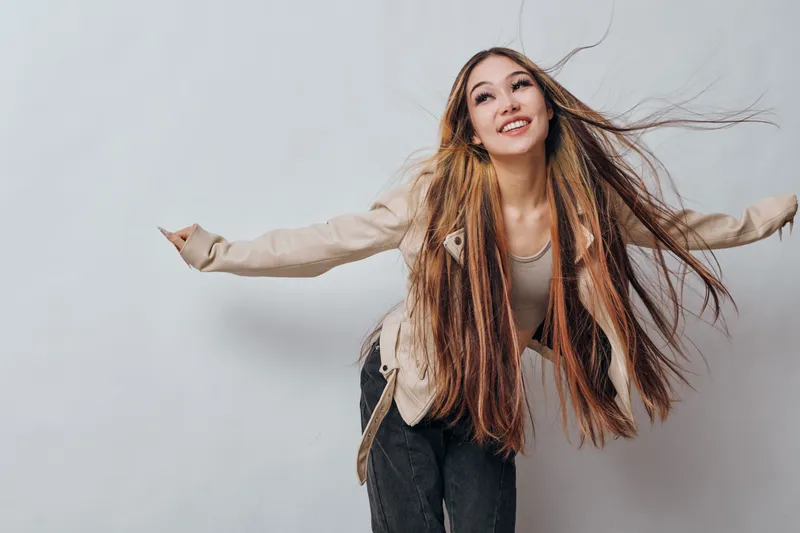 Young woman with long hair posing energetically against white background. Casual outfit with beige jacket and black pants. Smile, motion, fun and carefree mood in studio.