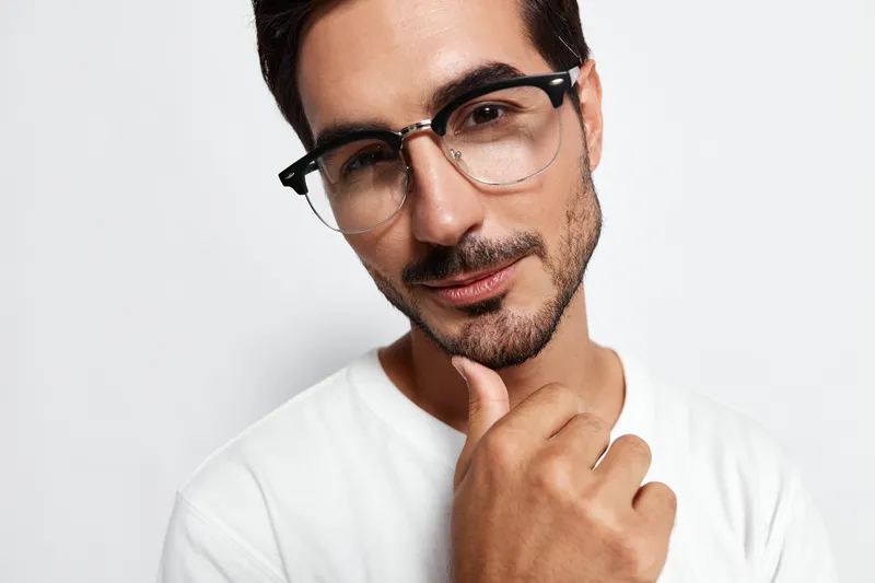 Stylish young man wearing glasses confidently poses for the camera, hand on chin, with a playful smile. Isolated on plain solid white background studio portrait.