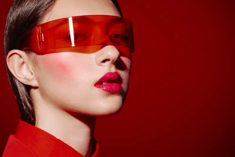 Stylish young woman in futuristic orange visor and red blazer with bold blush and glossy pink lips on red background. Creative beauty and fashion concept with vivid colors.