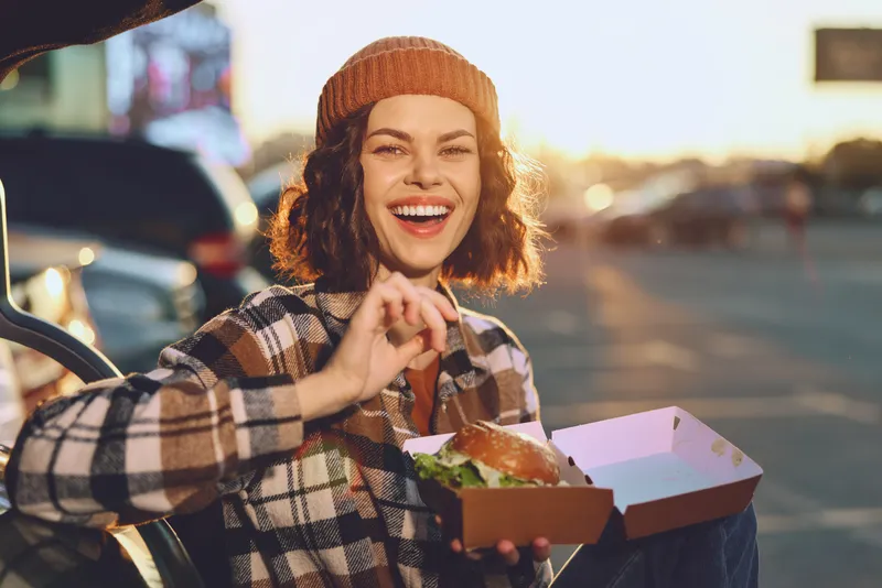 Woman with smile holding burger in takeaway box during golden hour in urban parking, candid portrait capturing authenticity, joyful lifestyle, relaxed evening sunlight and storytelling mood
