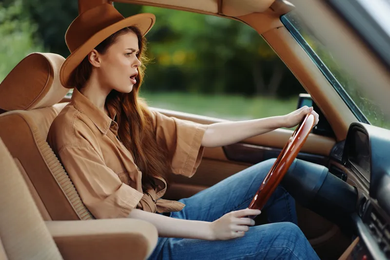 Stylish woman driving a classic car, showcasing adventurous spirit and confidence in a natural green setting with focused emotions.