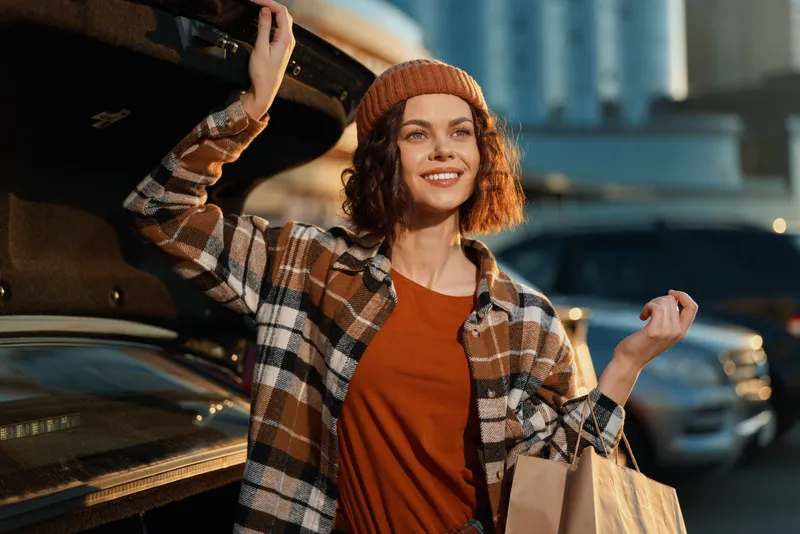 Woman smiling by open car trunk holding groceries in urban parking, wearing beanie and plaid jacket, candid lifestyle portrait with golden hour glow, authenticity and mindful living vibe.