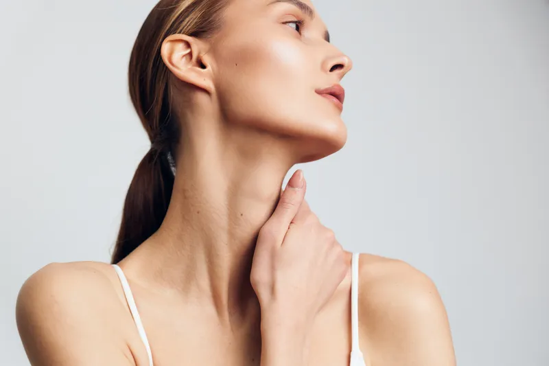 Young woman with long hair touches her neck gently while looking sideways against a plain light background, expressing calmness and natural beauty.