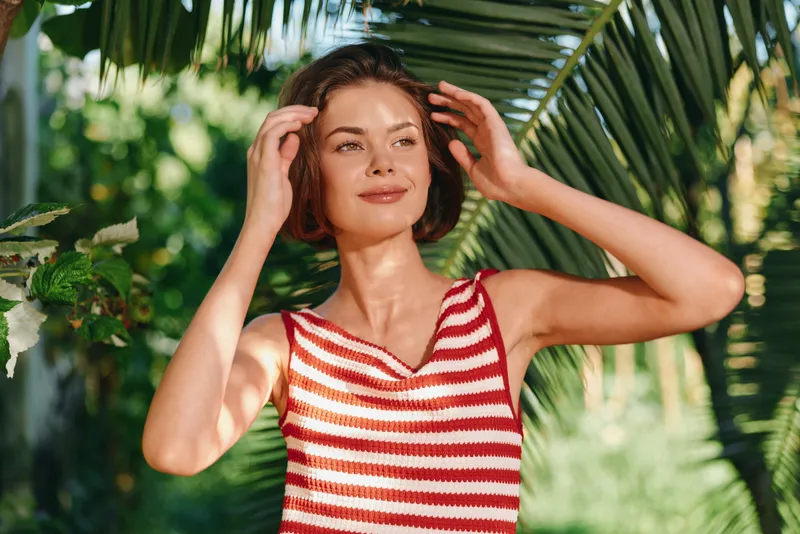 A cheerful woman wearing a red and white striped sleeveless top stands in a lush garden, smiling softly as she touches her hair, creating a warm, relaxed outdoor mood.