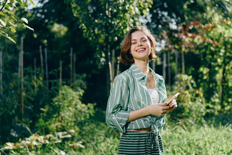 A smiling woman stands in a sunlit park, wearing a striped shirt and casual outfit, holding a phone.
