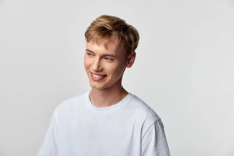 Smiling man in a casual white tee stands in a bright studio, conveying friendly energy and approachable style for lifestyle or fashion use with clean composition