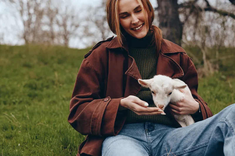 Young woman in a brown jacket gently feeds a white baby goat while sitting on grass in an outdoor natural setting during autumn.
