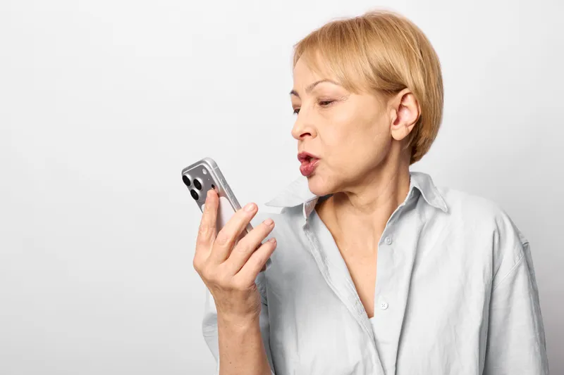 Older woman portrait, surprised shouting while holding phone, gaze fixed on screen, tense facial expression, communication and technology concept, isolated on white background