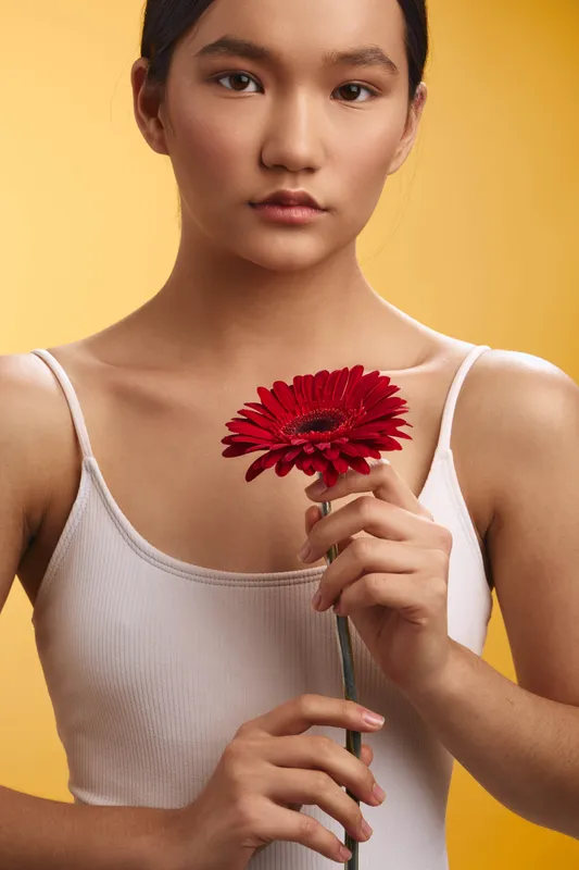 Asian woman looks directly at the camera while holding a red flower in front of a warm yellow background, creating a calm, graceful portrait with subtle confidence and gentle mood.