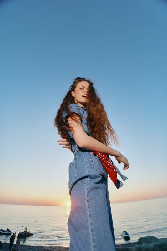lifestyle woman in modern boho-western denim street style standing by the sea at sunset with film color effect, casual and natural vibe