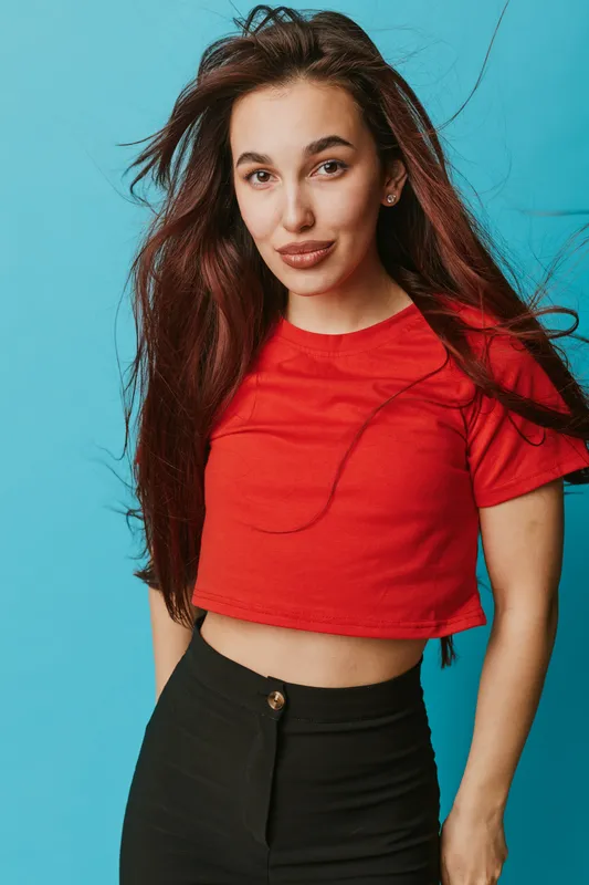 Young woman with long hair wearing red cropped t shirt and black pants posing against blue background. Casual style, confident expression, fashion portrait with natural look and loose hair.