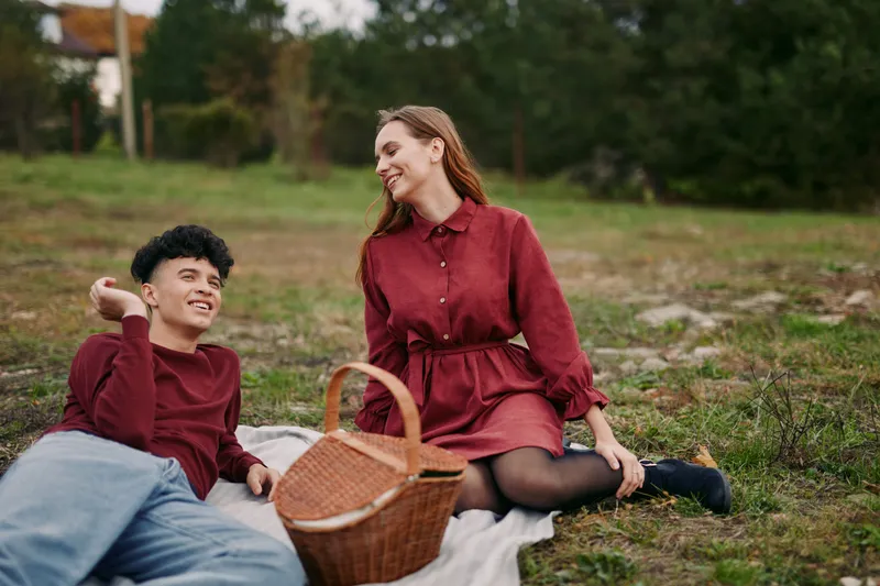A candid outdoor portrait of a young couple sharing a relaxed picnic on a grassy meadow, showing authenticity and credible natural connection with gentle smiles and warm body language.