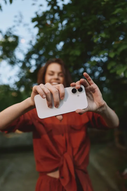 Cheerful woman outdoors taking a selfie, smiling at the camera, sunlit moment, casual red outfit, relaxed pose, natural colors, leafy background, lifestyle portrait during a warm afternoon