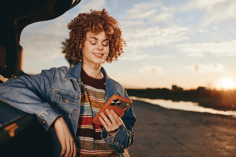 Woman lifestyle on seaside beach at sunset, curly hair and denim jacket, smiling while holding smartphone in her hands, casual outdoor travel mood with warm natural light and gentle sea breeze