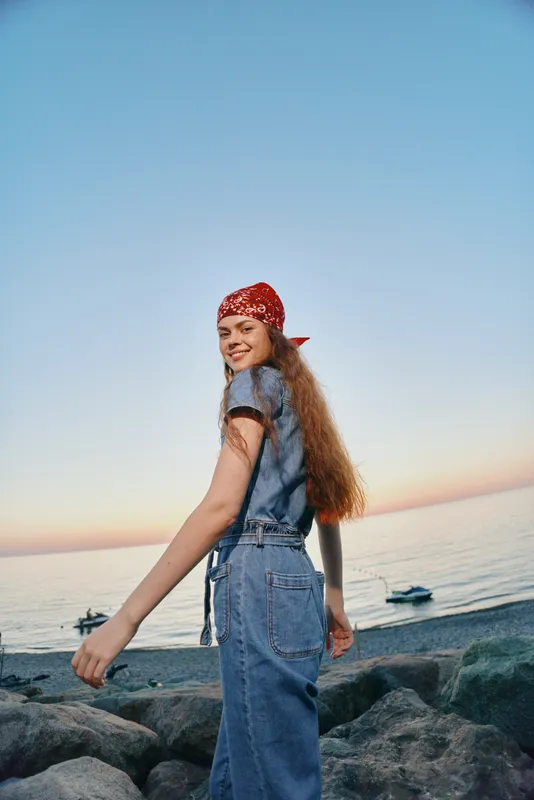 lifestyle woman in modern boho-western denim street style with red bandana smiling by the beach at sunset in warm film color tones