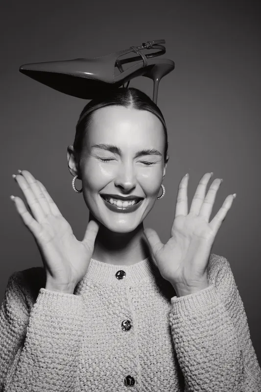 Young white woman with light skin smiling with closed eyes wearing earrings and a textured sweater holds her hands raised with a shoe balanced on her head in black and white.