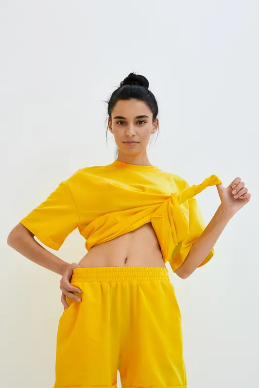 Young woman wearing a bright yellow outfit with a tied shirt and shorts poses confidently against a light background, showcasing casual style and sporty vibes.