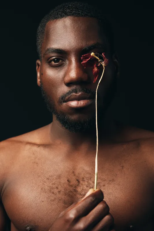 African man with dark skin holding dried flower in front of face, creative conceptual portrait with visual style, bare chest and serious expression on black background.