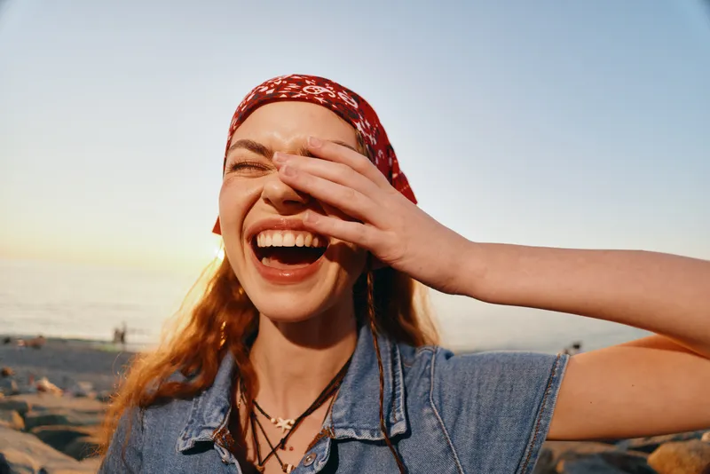 Lifestyle woman in modern boho-western denim street style laughs joyfully under warm sunlight with film color effect enhancing her vibrant carefree mood by the beach.