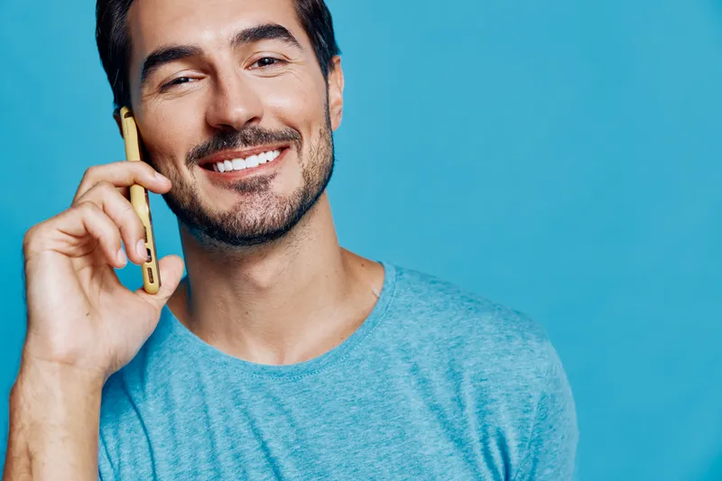 Smiling man with a cheerful expression, holding a mobile phone to his ear, wearing a light blue t-shirt against a vibrant blue background in a studio setting.
