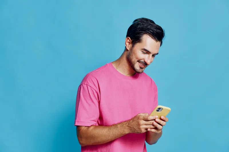 Smiling man wearing pink t-shirt looks at mobile phone with cheerful expression against vibrant blue background. Modern lifestyle concept in portrait style.