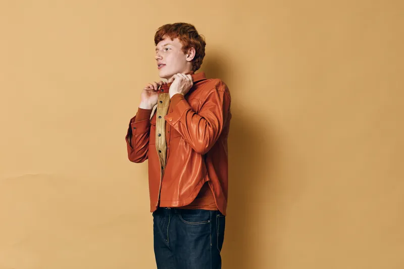 Young man with red hair wearing orange jacket and brown shirt standing against beige background, adjusting collar and looking sideways with thoughtful expression in casual outfit.