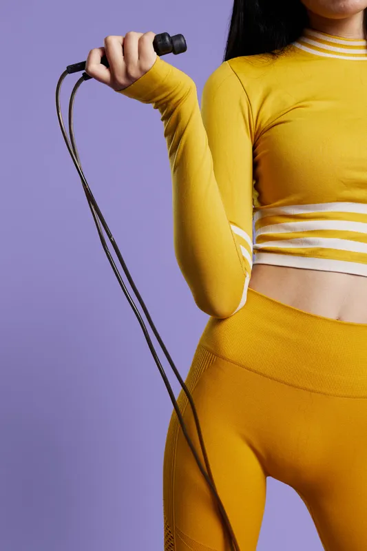 Woman sport fitness wearing yellow activewear holding a jump rope, demonstrating strength and readiness for workout with vibrant purple background.